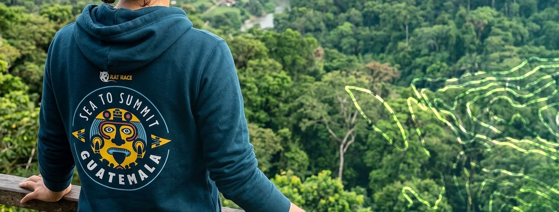 Person wearing a hoodie with a Guatemalan design, standing on a wooden platform overlooking a lush green landscape.